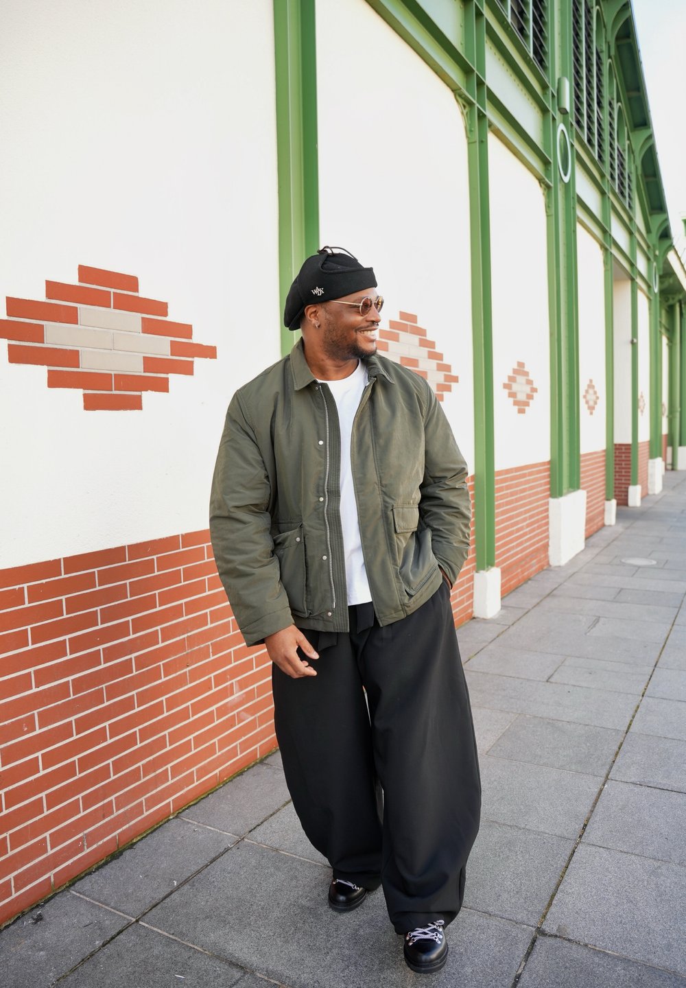 Homme souriant marchant le long d'un mur en briques blanches et vertes avec des poutres vertes, portant une veste verte, un pantalon large noir, un béret et des lunettes de soleil.