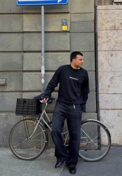 Camisa negra de manga larga con texto blanco, vaqueros negros holgados y zapatos negros brillantes al lado de una bicicleta plateada con una cesta negra apoyada contra una pared texturizada.