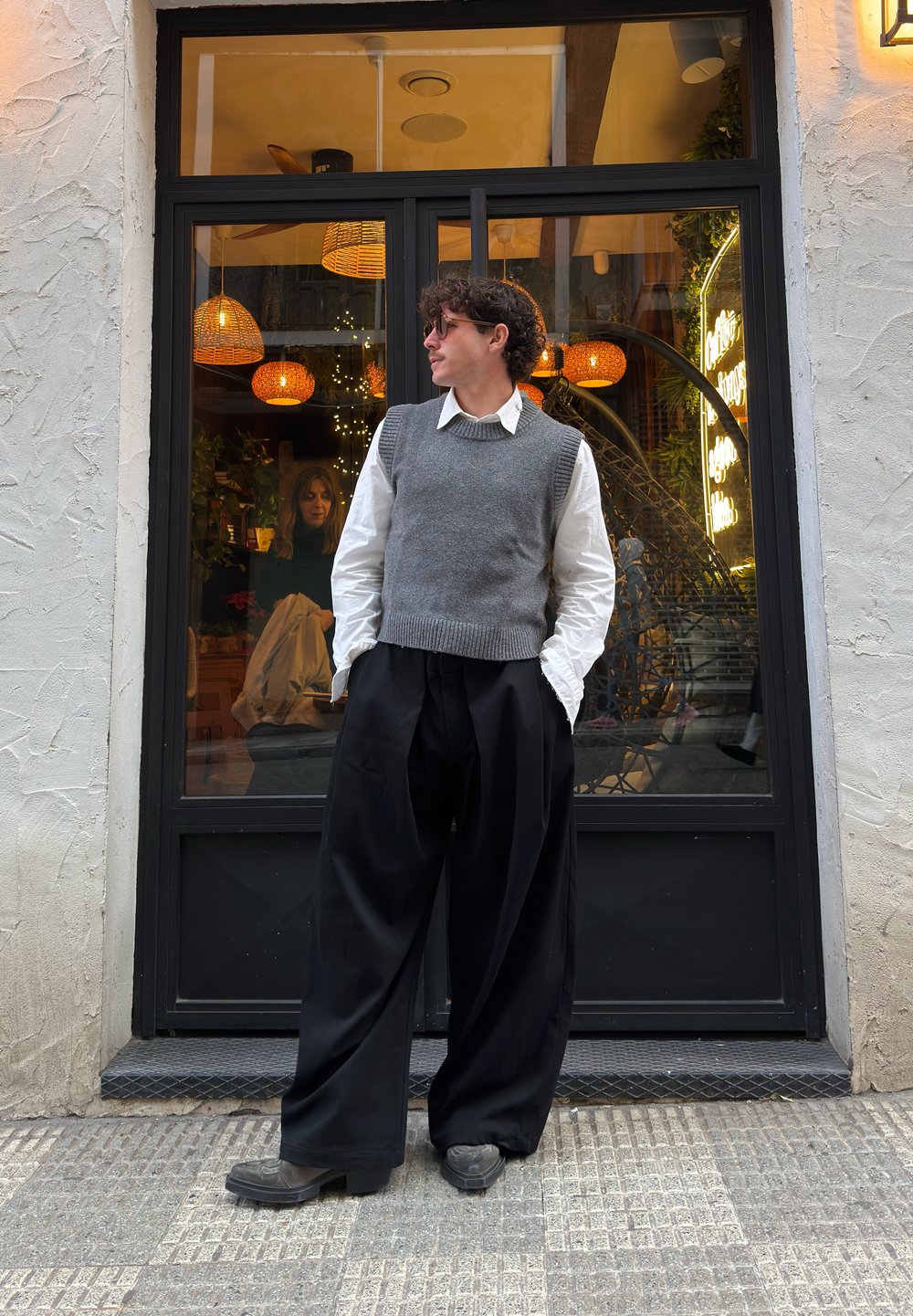 Gilet en tricot gris porté sur une chemise blanche à col, associé à un pantalon noir ample et à des chaussures foncées. En arrière-plan, des luminaires suspendus décoratifs.