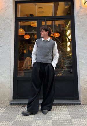 Chaleco de punto gris sobre una camisa blanca con cuello, combinado con pantalones negros holgados y zapatos oscuros. El fondo presenta lámparas colgantes decorativas.