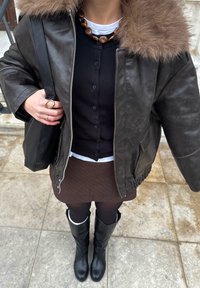 Cazadora bomber de cuero marrón con cuello de piel, suéter negro con botones, falda marrón con finas rayas, botas negras hasta la rodilla y bolso tote negro.