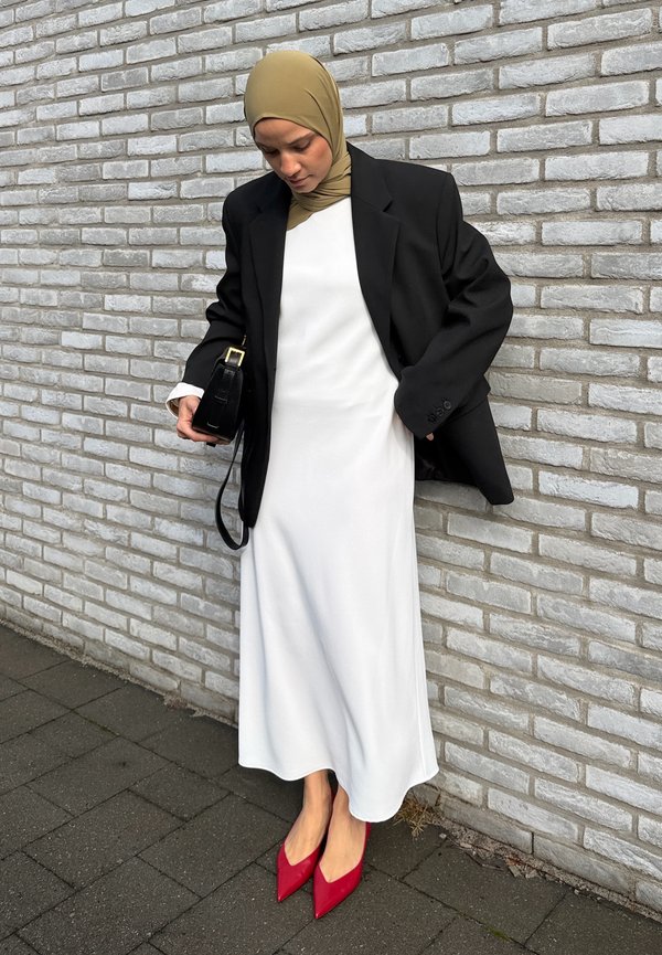 Une femme portant un hijab beige, un blazer noir, une robe blanche et des talons rouges tient un petit sac à main noir près d’un mur de briques.
