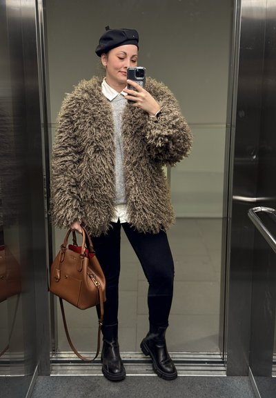 Mujer con boina negra, abrigo marrón grande y peludo, suéter gris sobre camisa blanca, pantalones negros, botas, sosteniendo bolso marrón, tomándose una selfie en el espejo del ascensor.