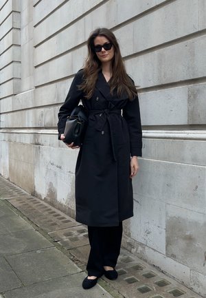 Abrigo negro de trench con cinturón, botones dobles y mangas largas, combinado con pantalones negros y zapatos planos contra una pared de piedra.