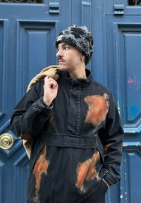 Black oversized denim jacket with orange paint splatters, high collar, and elastic cuffs. Layered over a light brown backpack. Knitted hat.