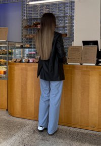 A person with long hair, wearing a black jacket and blue trousers, stands at a bakery counter where pastries and menus are displayed behind glass.