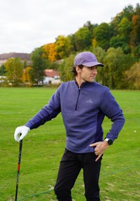 Lilla lukuga golfisärk tekstuursete triipudega, mustad püksid, valge kindaga, hoides golfikeppi. Mütsi peal on logo; taustal roheline muru.