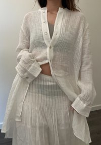 Camisa blanca de lino con un frente abotonado, mangas largas y un patrón texturizado. Llevada sobre una falda blanca plisada a juego.
