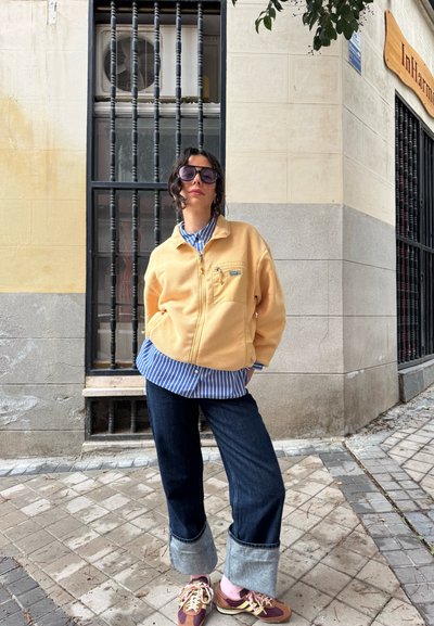 Chaqueta de forro polar con cremallera en amarillo pastel, camiseta a rayas debajo, jeans de mezclilla oscura con dobladillos remangados y zapatillas burdeos.