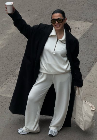 Mujer con chándal blanco y abrigo negro, sosteniendo un vaso de café para llevar en una mano y un gran bolso blanco en la otra, sonriendo al aire libre.
