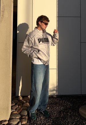 Rompevientos plateado de PUMA con un gran logo, combinado con unos jeans holgados de color azul claro y unas zapatillas de deporte verde oscuro, de pie contra una pared clara.