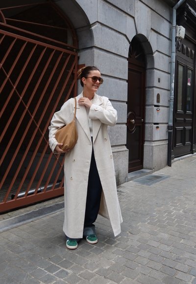 Abrigo largo color crema con grandes botones, usado sobre un suéter claro y pantalones oscuros. Zapatillas verdes y un bolso color marrón completan el look.