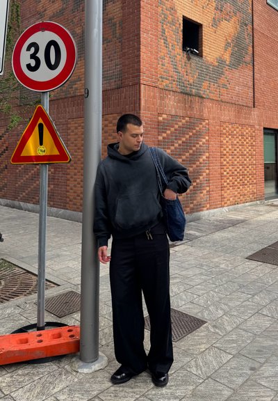 Sudadera negra, pantalones anchos negros y zapatos negros. Una bolsa azul cuelga del hombro. Entorno urbano con señales de tráfico y una pared de ladrillo.