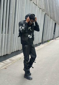 Fekete bőrkabát kapucnival, vaskos fekete pulóver fehér mintákkal, bőr nadrág és fekete fűzős bakancsok pántokkal és fém kiegészítőkkel.