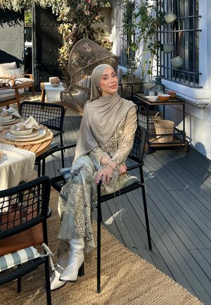 Persona sentada que lleva un hijab beige y un atuendo fluido con un patrón en colores claros. Botas blancas de tacón grueso. Ambiente de comedor al aire libre.