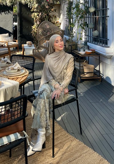 Persona sentada que lleva un hijab beige y un atuendo fluido con un patrón en colores claros. Botas blancas de tacón grueso. Ambiente de comedor al aire libre.