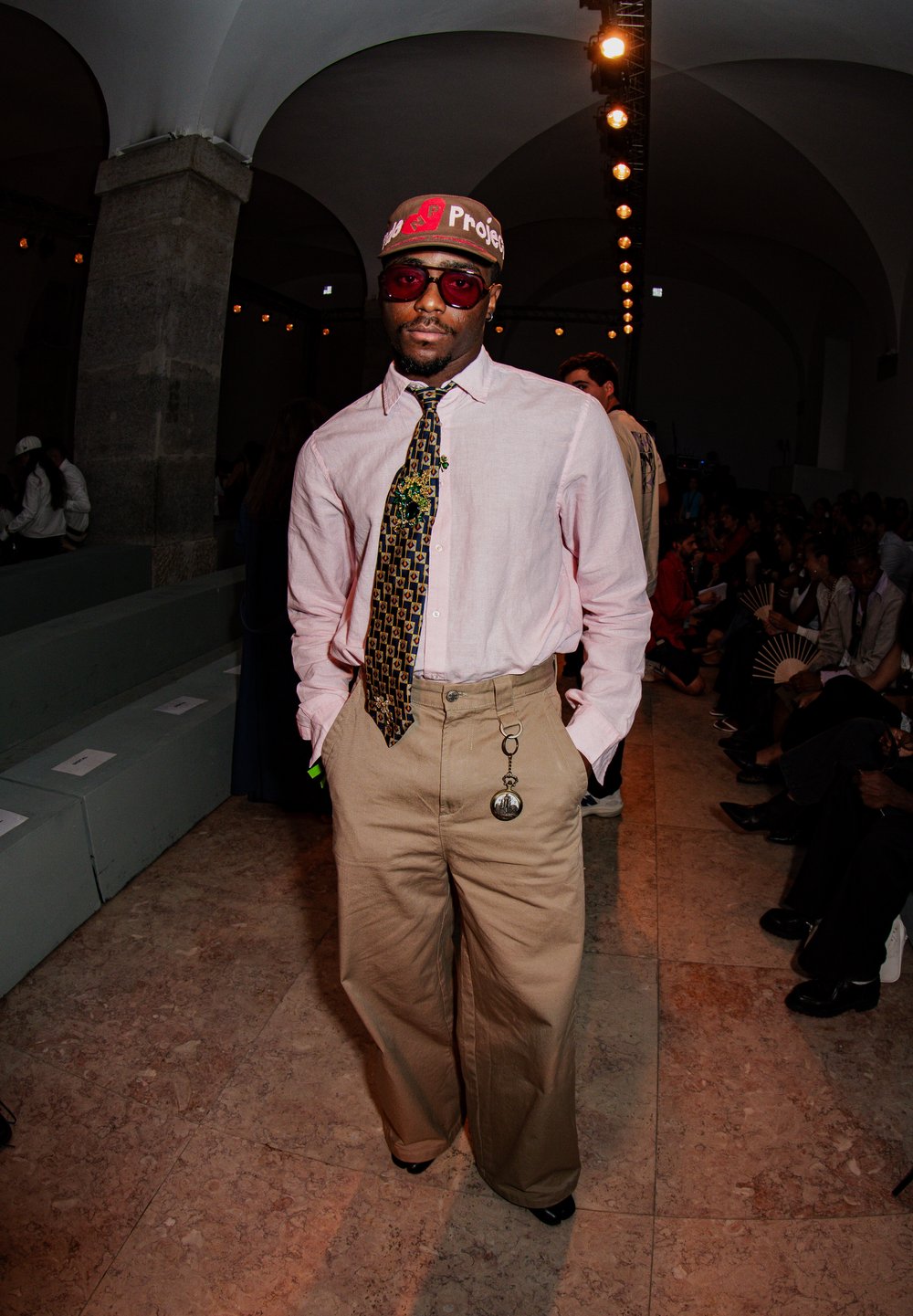 Beige wide-leg trousers paired with a pocket watch, a light pink button-up shirt, a patterned tie, and a red cap featuring colourful text. Black sunglasses complete the look.