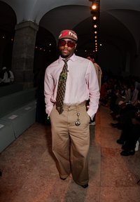 Beige wide-leg trousers paired with a pocket watch, a light pink button-up shirt, a patterned tie, and a red cap featuring colourful text. Black sunglasses complete the look.