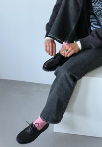 Chaussures en cuir noir à lacets, bout texturé, associées à des chaussettes rose clair. Un pantalon en denim gris et un pull foncé complètent la tenue.