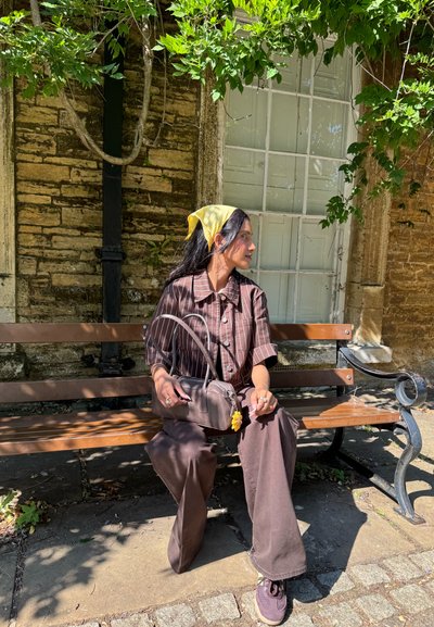 Mujer vestida con un atuendo marrón y un pañuelo amarillo se sienta en un banco de madera sosteniendo una bolsa, al lado de una pared de piedra con una gran ventana y vides verdes.