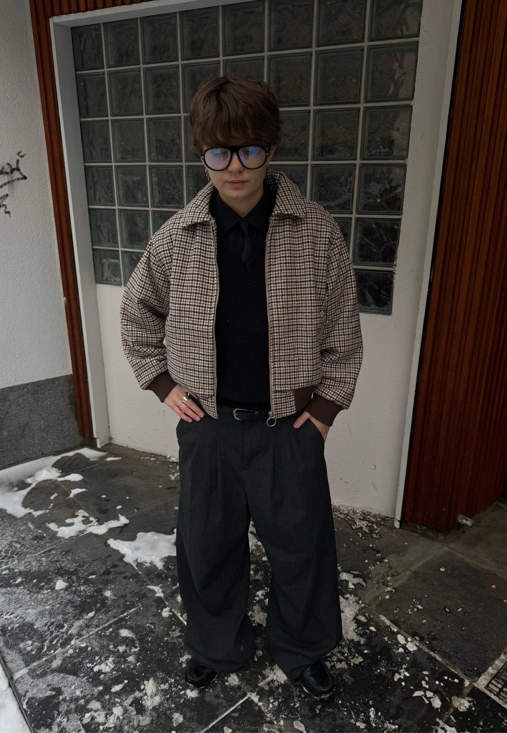 Veste courte marron à motif pied-de-poule, chemise noire, pantalon gris surdimensionné, chaussures noires, avec un mur en blocs de verre géométriques en arrière-plan.