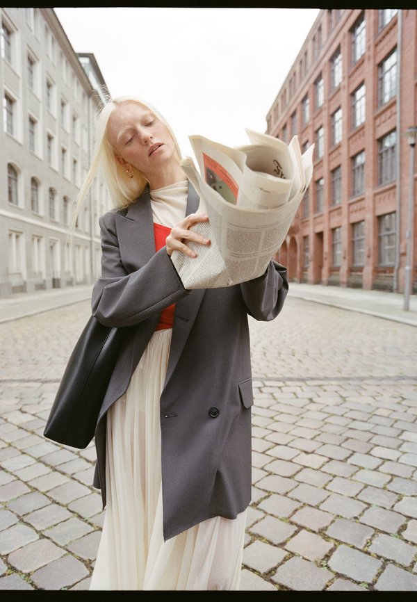 Szürke blézer fehér, pliszírozott ruhán, újságokat tart a kezében. Hosszú szőke haj, minimalista fekete táska. Macskaköves utca téglaépületekkel.
