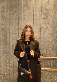 Sac à main noir avec ferrures circulaires dorées, manteau vert olive et colliers superposés. Mur texturé et rampe en bois en arrière-plan.