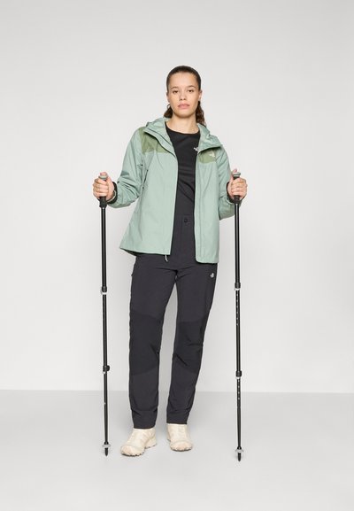 Chaqueta impermeable verde claro con capucha, camiseta negra, pantalones de senderismo negros y zapatos beiges, sosteniendo bastones de trekking.