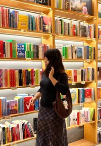 Woman browsing books on well-lit wooden shelves in a bookstore, wearing a black top, plaid skirt, and carrying a brown bag with a white plush keychain.