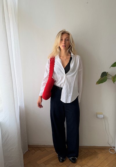 Mujer con el pelo largo y rubio lleva una camisa blanca, pantalones negros, zapatos negros y un bolso rojo al hombro, de pie en un interior cerca de una planta y una ventana.
