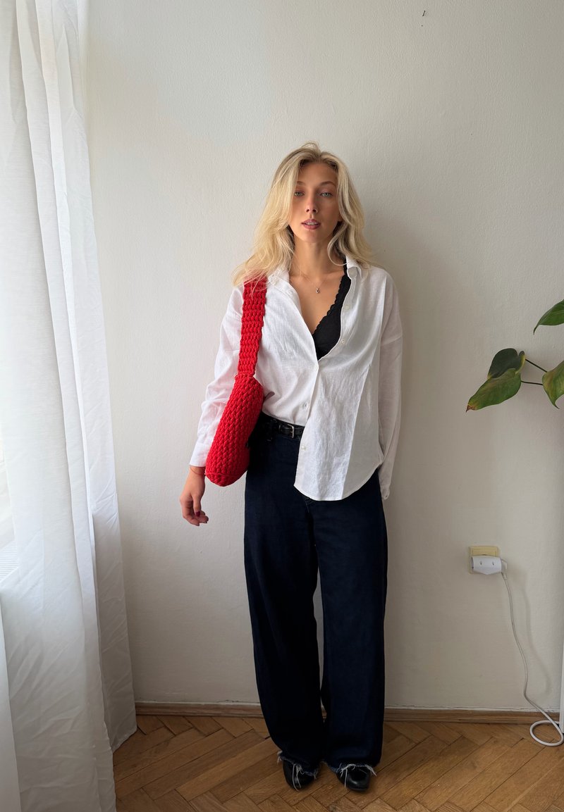 Femme aux longs cheveux blonds porte une chemise blanche, un pantalon noir, des chaussures noires et un sac à bandoulière rouge, debout à l’intérieur près d’une plante et d’une fenêtre.