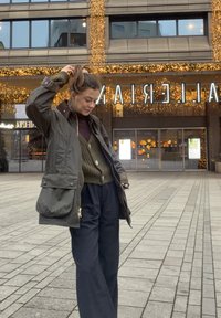 Donkergroene, waterafstotende jas met een grote voorzak, gedragen over een bruine gebreide trui en marineblauwe wijde broek, buiten gedragen.