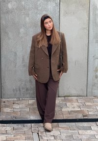 Blazer marrone oversize con doppi bottoni sopra una camicia scura e pantaloni marroni, abbinati a stivaletti beige e fascia nera per capelli.