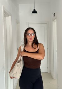 Brun ärmlös topp, högmidjade svarta leggings och en texturerad krämfärgad handväska. Modern inredning med vita väggar och en svart hängande lampa.
