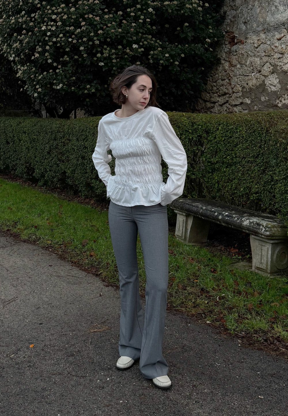 Λευκή μπλούζα με μακριά μανίκια και στραμμένα λεπτομέρειες στη μέση, συνδυασμένη με ανοιχτό γκρι φαρδύ παντελόνι και κρέμα παπούτσια. Εξωτερικός χώρος.