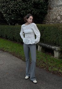 Λευκή μπλούζα με μακριά μανίκια και στραμμένα λεπτομέρειες στη μέση, συνδυασμένη με ανοιχτό γκρι φαρδύ παντελόνι και κρέμα παπούτσια. Εξωτερικός χώρος.