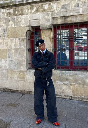 Veste en denim foncé avec un col, pantalon en denim foncé ample, béret noir et chaussures orange vif, le tout devant un mur en pierre.