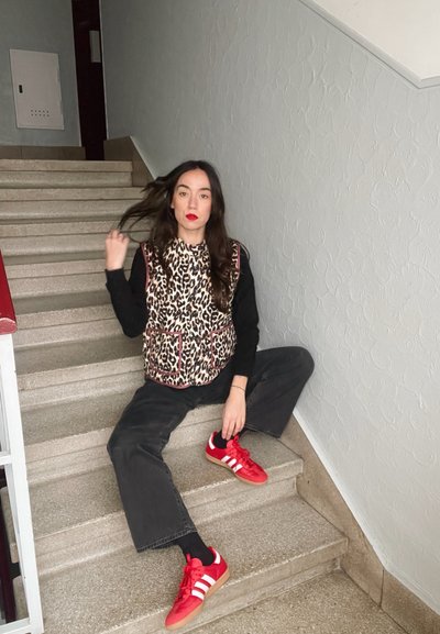 Chaleco de estampado de leopardo con acentos rojos sobre una camiseta de manga larga negra, combinado con jeans oscuros y zapatillas deportivas rojas con franjas blancas.