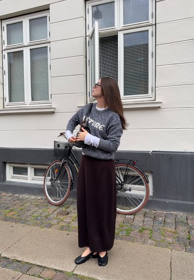Suéter gris de punto con el texto "Futuro", camiseta blanca debajo, falda larga marrón, zapatos negros, de pie junto a una bicicleta contra una pared con ventanas.