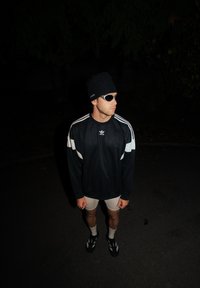 Černá sportovní mikina s bílými pruhy, světle šedé šortky, černé ponožky, černé sandály, černá čepice a stříbrné sluneční brýle.