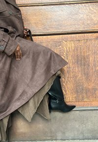 Brun semsket trenchcoat iført over et beige plagg, kombinert med svarte spisse støvler med spenne. Trebakgrunn.