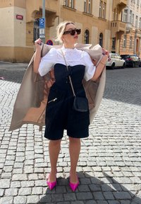 Top negro sin tirantes con detalle de lazo, blusa blanca de eyelet, pantalones cortos negros y tacones altos rosas. Lleva un bolso cruzado negro y un abrigo beige.