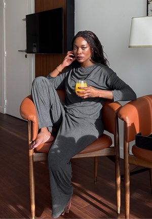 Conjunto de ropa cómoda de manga larga gris hecho de tela suave; sentado en una silla de cuero color canela, sosteniendo un vaso con bebida naranja, con los pies descalzos.