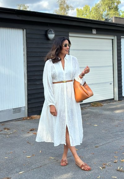 Vestido blanco de manga larga con tejido texturizado, ceñido en la cintura. Cinturón marrón, sandalias a juego y un bolso naranja. Entorno urbano al aire libre.