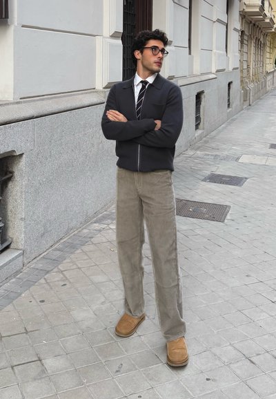Chaqueta oscura con cremallera sobre una camisa blanca y una corbata de rayas; pantalones de pana beige claro; botines marrones; de pie en la acera de una ciudad.