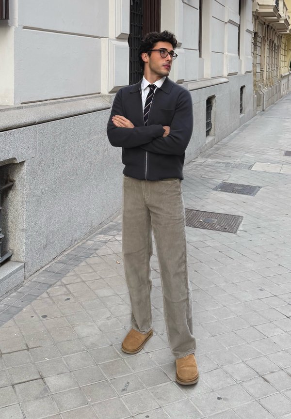 Veste zippée sombre par-dessus une chemise blanche et une cravate rayée ; pantalon en velours côtelé beige clair ; bottines marron ; debout sur un trottoir en ville.