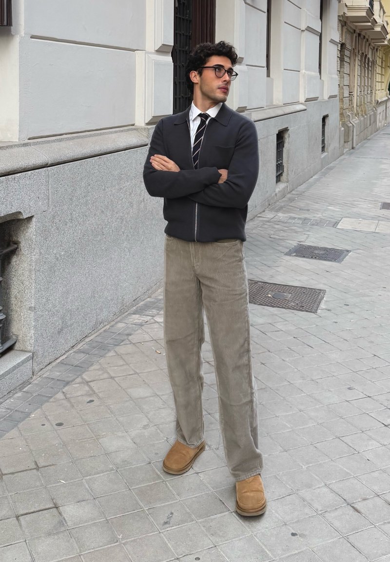 Veste zippée sombre sur une chemise blanche et une cravate rayée ; pantalon en velours côtelé beige clair ; bottines marron ; debout sur un trottoir en ville.