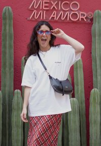 T-shirt oversized branca com o texto "ALLSAINT", saia xadrez vermelha, bolsa crossbody preta com tachas. O fundo apresenta cactos verdes e uma parede vermelha.