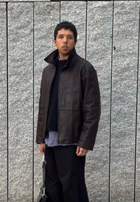 Καφέ δερμάτινο μπουφάν με ψηλό γιακά, φορεμένο πάνω από ένα υφασμάτινο μαύρο πουλόβερ και καρό πουκάμισο; μαύρο παντελόνι και κρατώντας μια τσάντα με ασημένιο κούμπωμα.