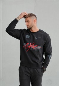 Sweat-shirt noir avec graphiques imprimés en rouge et gris. Dispose d'un col rond, de poignets côtelés et d'un design texte unique avec des noms de villes.